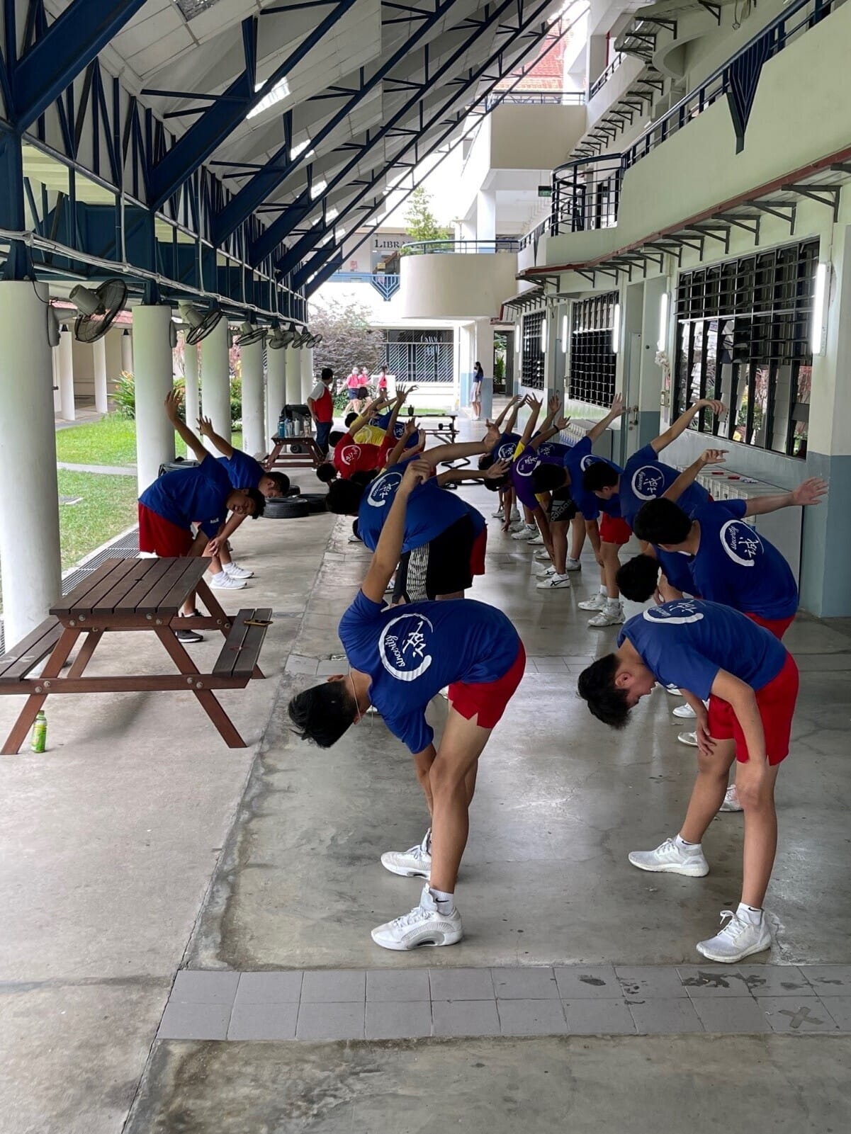 A group of students in blue shirts and red shorts stretch sideways in a covered school corridor.